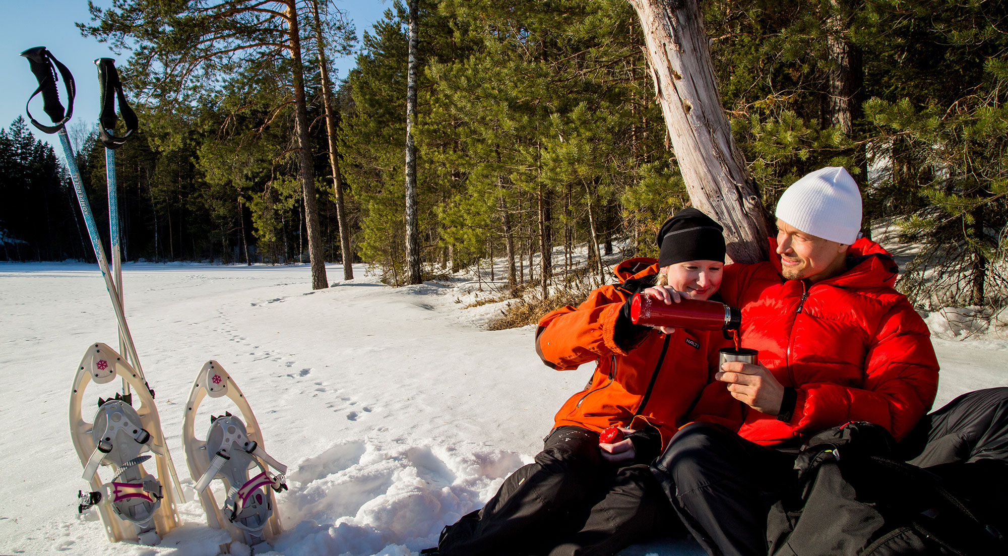 Punaisiin takkeihin pukeutuneete mies ja nainen istuvat lumisen lammen rannalla. Lumikengät on riisuttu viereen. Termospullosta kaadetaan kuumaa juotavaa kuksaan.
